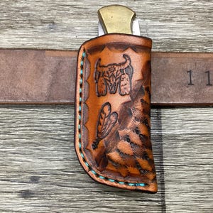May include: A tan leather knife sheath featuring a stamped steer skull and feather design with turquoise stitching. The sheath is attached to a brown leather belt clearly marked with the measurement 1 1/2 inches, holding a folding knife with a brass bolster.