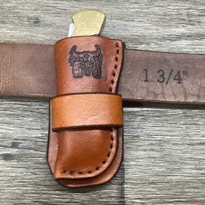 May include: A tan leather knife sheath with a stamped bull skull design, featuring a horizontal belt loop attachment. The sheath is shown mounted on a brown leather belt with the text 1 3/4" printed on the surface, indicating the belt width.