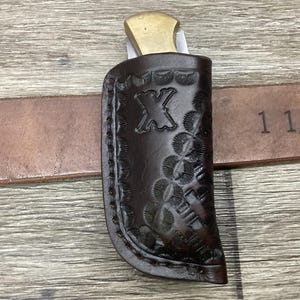 May include: A dark brown leather knife sheath with a stamped floral border and the letter X embossed on the front. The sheath holds a folding knife with a brass bolster and is positioned on a brown leather belt marked with 1 1/2 inches.
