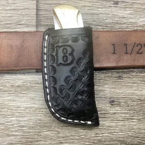 May include: A black leather knife sheath with a stamped letter B and a basket weave pattern, featuring white stitching along the edge. The sheath holds a folding knife with a brass bolster and is positioned on a brown 1 1/2 inch belt.