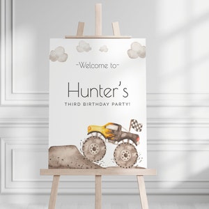 May include: A watercolor-style sign on a wooden easel. The sign reads "Welcome to Hunter's Third Birthday Party!" and features a yellow and black monster truck with flames and a checkered flag, with brown clouds.