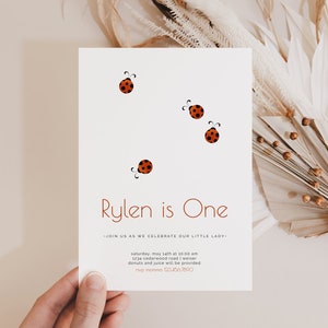 May include: A white card with red ladybugs and the text "Rylen is One" in brown. The card also includes the text "Join us as we celebrate our little lady" and details for a party.