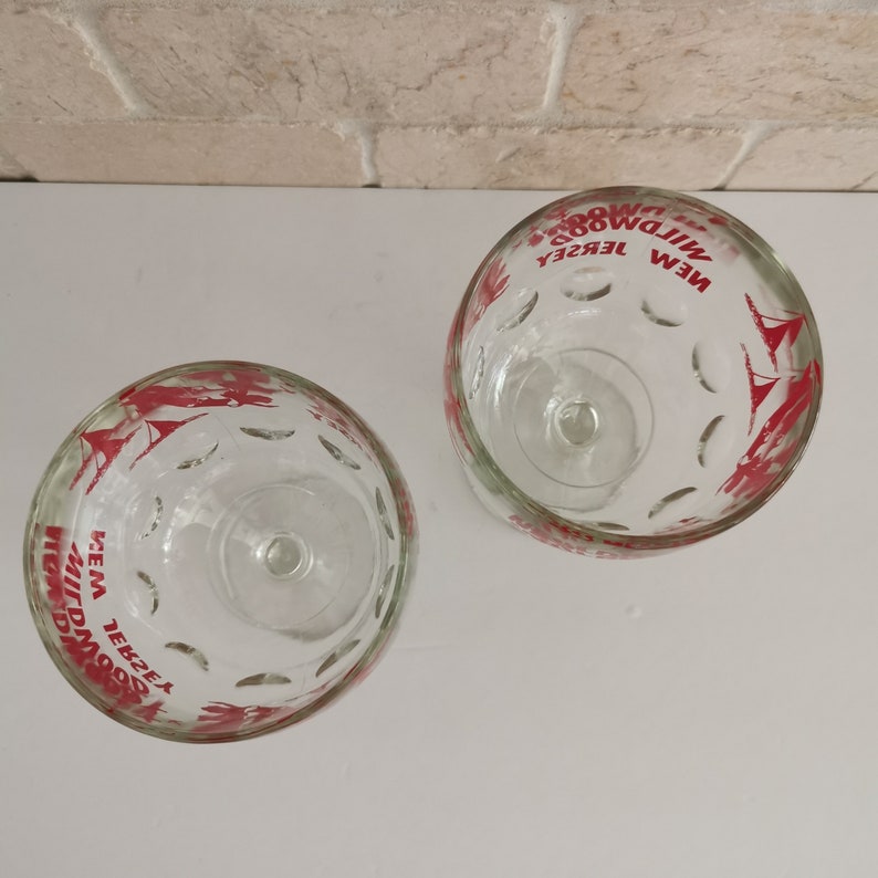 May include: Two clear glass cups with red printed designs. The cups have a scalloped rim and a small stem. The text on the cups reads "NEW JERSEY WILDWOOD" and "NEW JERSEY WILDWOOD KEM", respectively.