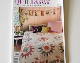 May include: A copy of "QUILTmania," The Quilt Magazine, featuring a colorful quilt with starburst patterns. The magazine cover displays the title and issue number. The background shows a bed with pillows and decorative items.