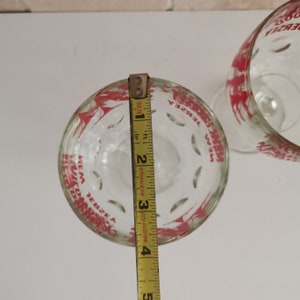 May include: Two clear glass wine glasses with red printed designs. The glasses have the words "New Jersey" printed on them. A measuring tape is used to measure the height of one of the glasses, which is 3.5 inches tall.