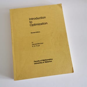 Puede incluir: Un libro amarillo titulado "Introducción a la optimización" con los nombres de los autores "Gerald Berman" y "K. D. Fryer" y el texto "Edición revisada" y "Facultad de Matemáticas Universidad de Waterloo".