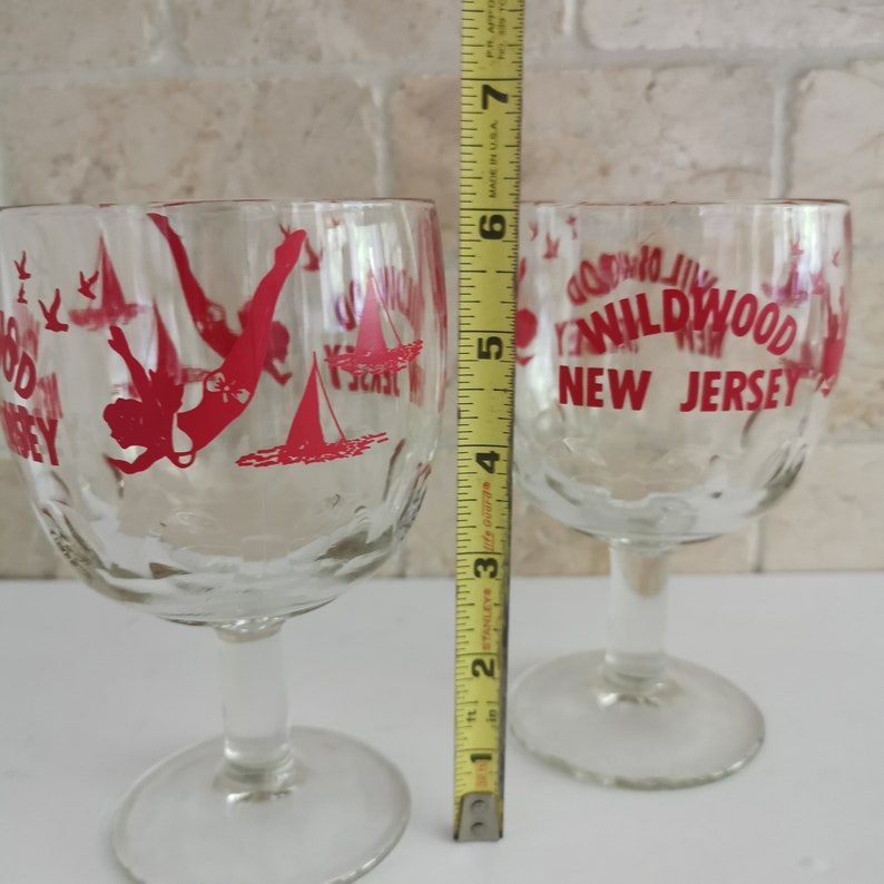 May include: Two clear glass wine glasses with red lettering that says "Wildwood New Jersey". One glass has a red silhouette of a woman diving into the water with a sailboat in the background.