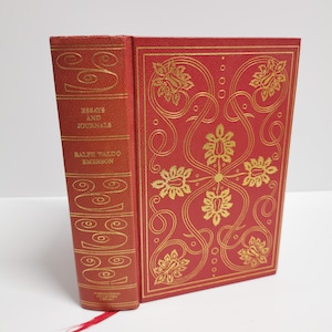 May include: A red hardcover book with gold lettering on the spine. The title is "Essays and Journals" by Ralph Waldo Emerson. The cover features a gold floral design.