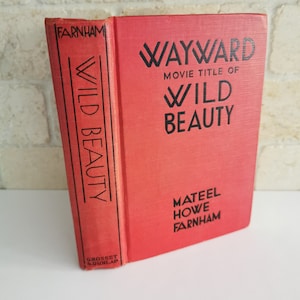 Puede incluir: Un libro de tapa dura rojo titulado "Wayward, Movie Title of Wild Beauty" de Mateel Howe Farnham. El libro está publicado por Grosset & Dunlap.