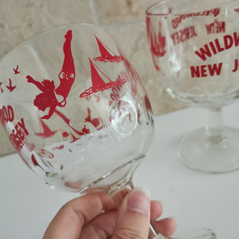 May include: Two clear glass goblets with red printed designs. One goblet features a woman diving into water with sailboats in the background. The other goblet has the text "WILDWOOD NEW JERSEY" printed on it.