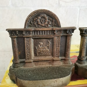 May include: A pair of antique cast iron bookends in the shape of a classical building with a decorative design. The bookends feature a relief sculpture of a person sitting in a chair.