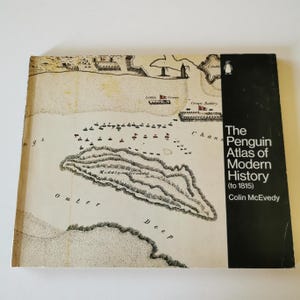 May include: The Penguin Atlas of Modern History (to 1815) by Colin McEvedy. The book cover features a detailed, vintage map illustration in shades of beige and brown, with a black spine. The map depicts a coastal area with ships and land formations.