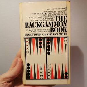 May include: The cover of a book titled "The Backgammon Book" by Oswald Jacoby and John R. Crawford. The book cover features a black and white backgammon board with red triangles.