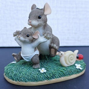 May include: A figurine featuring a large grey mouse with a white flower on its head standing behind a smaller grey mouse wearing a white nappy. The mice are positioned on a patch of green grass with white flowers, a cream snail, and a red ladybird.