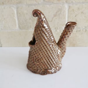 May include: A ceramic teapot with a textured, brown and gold glaze. The teapot has a unique, whimsical shape with a curved spout and a tall, pointed lid.