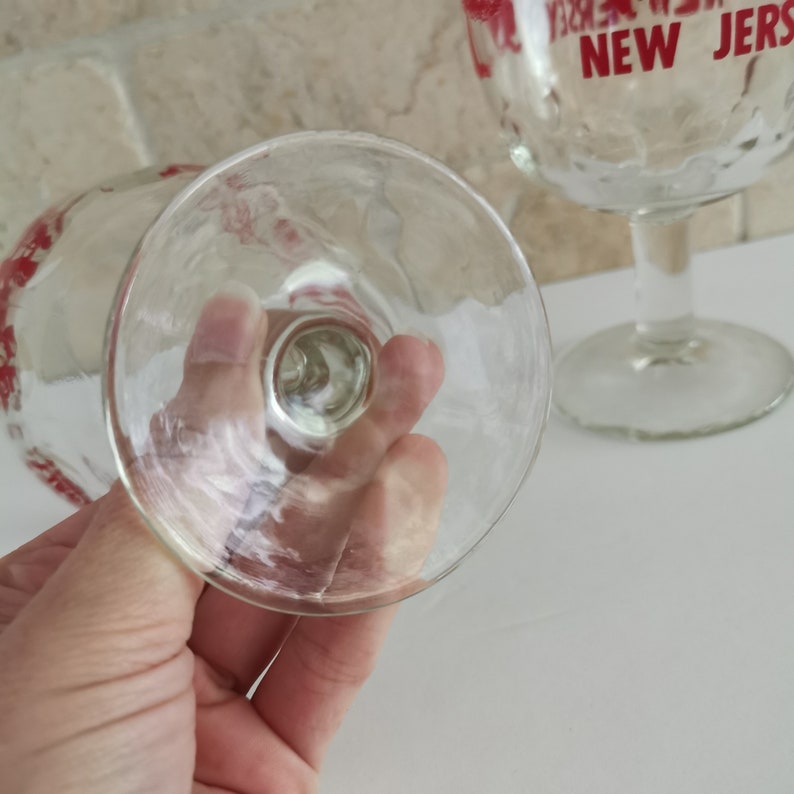 May include: Two clear glass goblets with red and white designs. The goblets have a wide bowl and a tall stem. One goblet has the words "NEW JERSEY" printed on it.