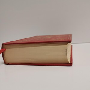 May include: A red hardcover book with a gold-colored design on the cover. The book is closed and the spine is visible. The book has a red ribbon bookmark.