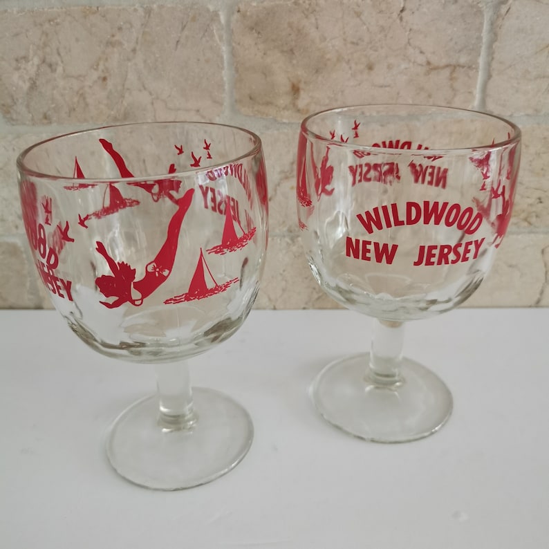 May include: Two clear glass goblets with red lettering that reads "Wildwood New Jersey". The goblets feature a silhouette of a woman diving into the water with birds and sailboats in the background.