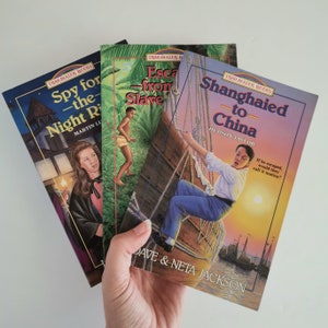 以下が含まれることがあります： 冒険と歴史小説に関連するカラフルな表紙とタイトルがついたペーパーバックの書籍が3冊あります。タイトルは「スパイ・フォー・ザ・ナイト・ライダーズ」、「エスケープ・フロム・スレイバリー」、「シャンハイド・トゥ・チャイナ」です。
