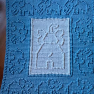 May include: A blue crocheted baby blanket with a white crocheted house design in the centre. The blanket has a textured, puffy design.