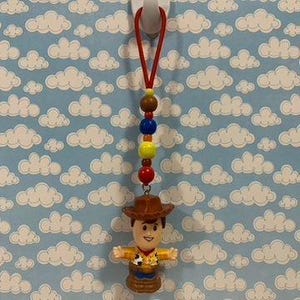 Puede incluir: Un dije de figura de acci&oacute;n de Woody con una cuerda roja, cuentas coloridas y un sombrero de vaquero marr&oacute;n. La figura tiene los brazos abiertos sobre un fondo azul con patrones de nubes blancas.