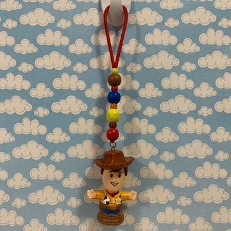 Puede incluir: Un dije de figura de acci&oacute;n de Woody con una cuerda roja, cuentas coloridas y un sombrero de vaquero marr&oacute;n. La figura tiene los brazos abiertos sobre un fondo azul con patrones de nubes blancas.