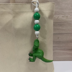 K&ouml;nnte beinhalten: Ein gr&uuml;ner Dinosaurier-Anh&auml;nger h&auml;ngt an einer wei&szlig;en Kordel mit gr&uuml;nen und wei&szlig;en Perlen. Der Dinosaurier hat eine leuchtend gr&uuml;ne Farbe und ist mit einem silberfarbenen Verschluss an der Kordel befestigt. Der Anh&auml;nger h&auml;ngt an einer beigen Tragetasche.