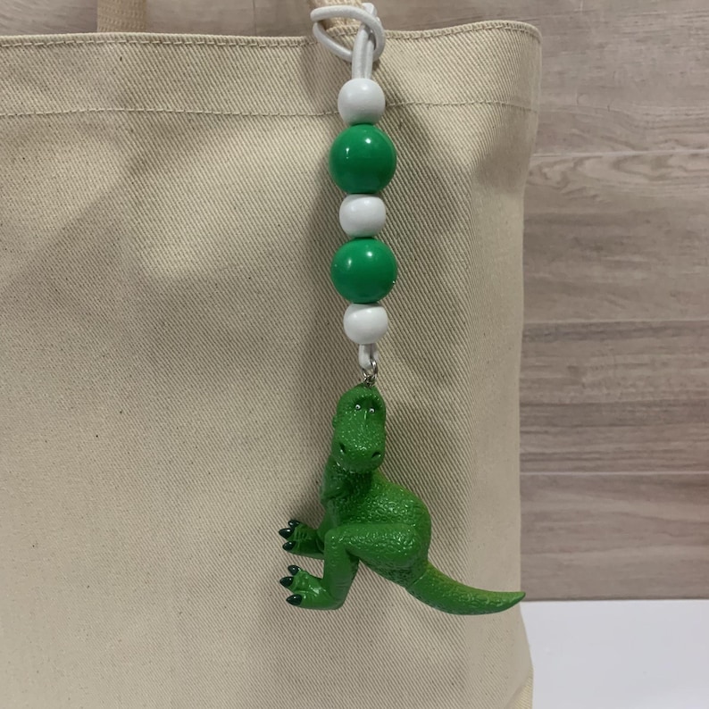 K&ouml;nnte beinhalten: Ein gr&uuml;ner Dinosaurier-Anh&auml;nger h&auml;ngt an einer wei&szlig;en Kordel mit gr&uuml;nen und wei&szlig;en Perlen. Der Dinosaurier hat eine leuchtend gr&uuml;ne Farbe und ist mit einem silberfarbenen Verschluss an der Kordel befestigt. Der Anh&auml;nger h&auml;ngt an einer beigen Tragetasche.