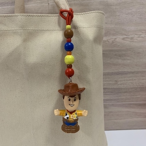 Puede incluir: Un amuleto de cuentas de colores con una figura de Woody de Toy Story. El amuleto tiene un cord&oacute;n rojo y una serie de cuentas en marr&oacute;n, azul, amarillo y rojo. La figura de Woody lleva un sombrero marr&oacute;n, una camisa amarilla y pantalones azules. El amuleto est&aacute; unido a un bolso tote beige.