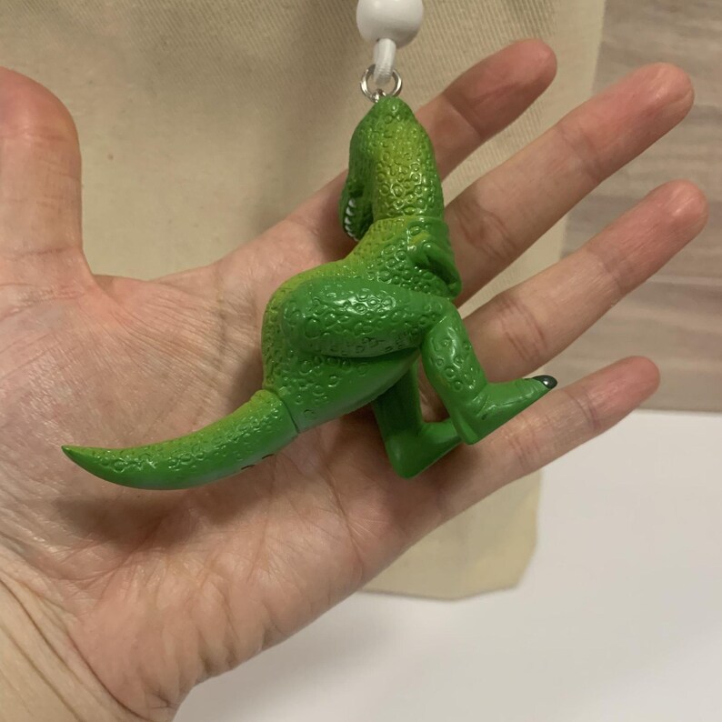 K&ouml;nnte beinhalten: Gr&uuml;ne Dinosaurierfigur mit strukturierter Oberfl&auml;che. Der Dinosaurier ist mit zur&uuml;ckgedrehtem Kopf und ausgestrecktem Schwanz posiert. Die Figur ist an einer wei&szlig;en Schnur und einem wei&szlig;en Ball befestigt.