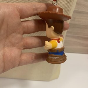 Puede incluir: Una figura de Woody de Little People, con sombrero de vaquero marr&oacute;n, camisa amarilla y vaqueros azules. La figura tiene un pa&ntilde;uelo rojo y est&aacute; sujeta a una cuerda roja. La base de la figura es marr&oacute;n y tiene la inscripci&oacute;n "Little People".