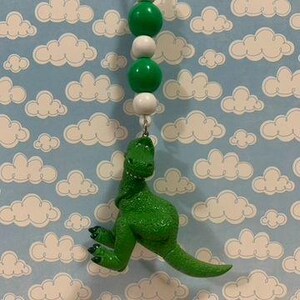 K&ouml;nnte beinhalten: Ein gr&uuml;ner Tyrannosaurus Rex-Anh&auml;nger h&auml;ngt an einer Perlenkette mit gr&uuml;nen und wei&szlig;en Perlen. Der Dinosaurier hat eine strukturierte Oberfl&auml;che und h&auml;ngt vor einem hellblauen Hintergrund mit wei&szlig;en Wolken.