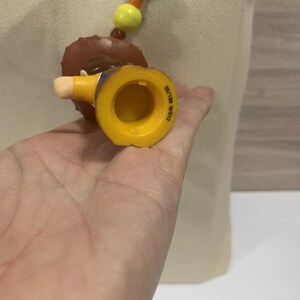 Puede incluir: Una figura de juguete amarilla y marr&oacute;n de un vaquero, con un sombrero marr&oacute;n y una camisa amarilla. La figura es parte de un collar con cuentas naranjas, amarillas y marrones. La figura se sostiene en una mano.