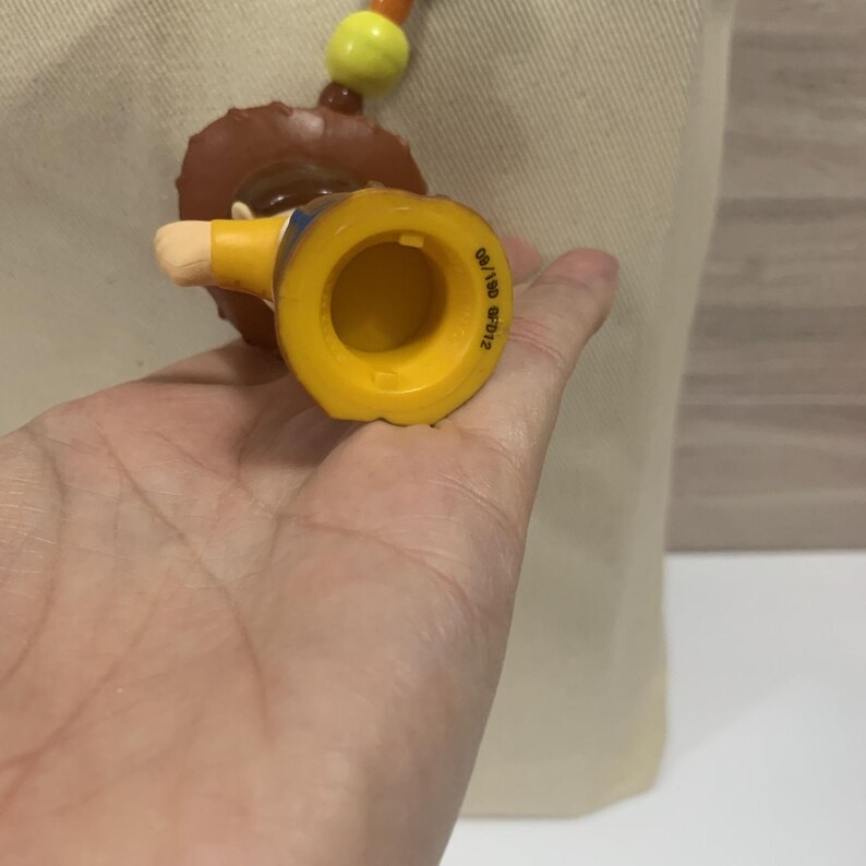 Puede incluir: Una figura de juguete amarilla y marr&oacute;n de un vaquero, con un sombrero marr&oacute;n y una camisa amarilla. La figura es parte de un collar con cuentas naranjas, amarillas y marrones. La figura se sostiene en una mano.