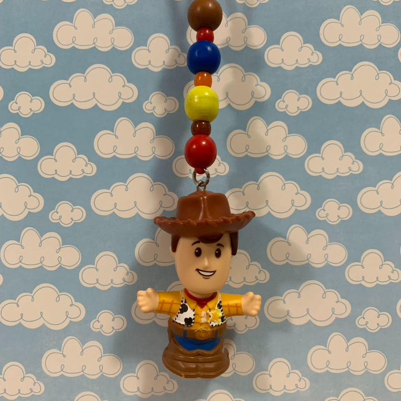 Puede incluir: Un dije de figura de Woody con sombrero de vaquero marr&oacute;n, camisa amarilla y vaqueros azules. El dije est&aacute; unido a una cadena de cuentas de colores. El fondo es un patr&oacute;n azul claro de nubes blancas.