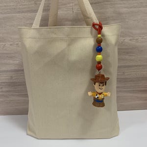 Puede incluir: Un bolso tote de lona beige con asas a juego. Un amuleto de cuentas de colores con una figura de Woody cuelga del asa. El amuleto tiene cuentas marrones, azules, amarillas y rojas.