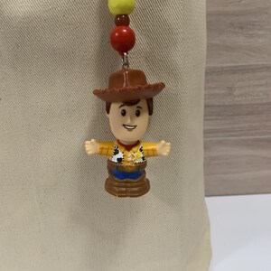 Puede incluir: Un dije de Woody con sombrero de vaquero marr&oacute;n, camisa amarilla y pantalones azules. El dije est&aacute; unido a una cadena de cuentas con cuentas rojas, marrones y amarillas. El dije cuelga de una tela beige.