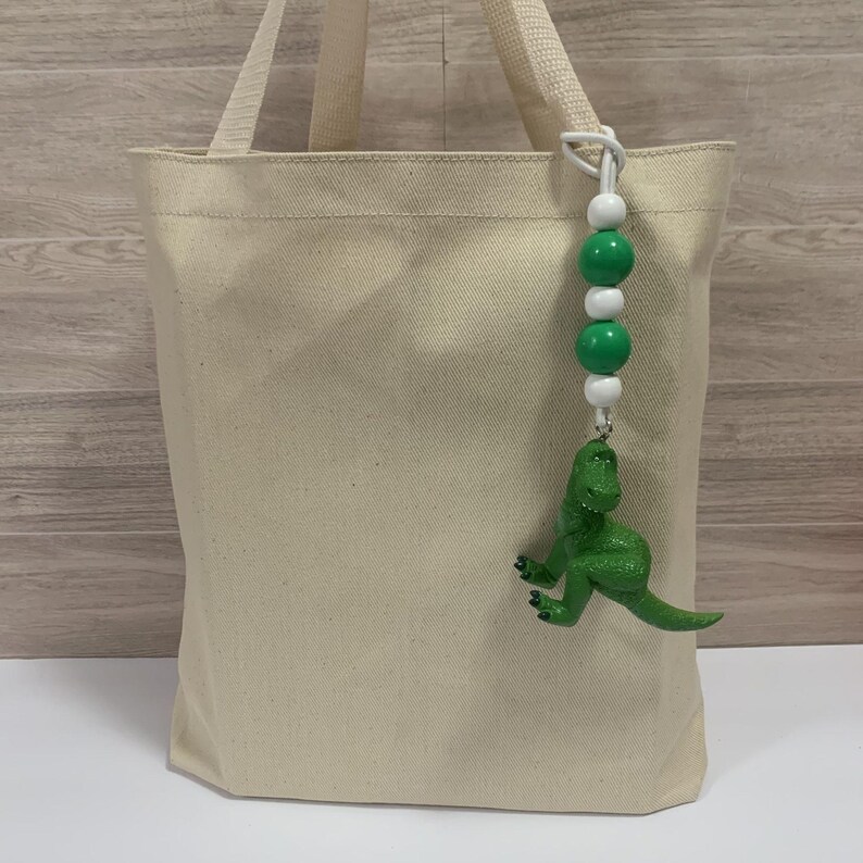 K&ouml;nnte beinhalten: Eine beigefarbene Canvas-Tragetasche mit passenden Griffen. Ein Perlenanh&auml;nger mit gr&uuml;nen und wei&szlig;en Perlen und einer gr&uuml;nen Dinosaurierfigur h&auml;ngt oben. Die Tasche ist ein einfacher, l&auml;ssiger Stil.
