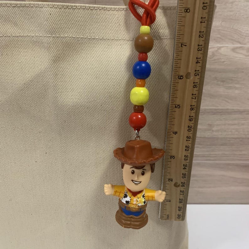 Puede incluir: Un llavero de cuentas de colores con una figura de Woody de Toy Story. El llavero tiene un cord&oacute;n rojo, cuentas marrones, azules, amarillas y rojas. La figura de Woody lleva un sombrero marr&oacute;n, una camisa amarilla y pantalones azules. El llavero mide unos 7.6 cm.