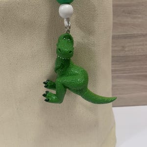 K&ouml;nnte beinhalten: Ein gr&uuml;ner Dinosaurier-Anh&auml;nger mit strukturierter Oberfl&auml;che, der an einer wei&szlig;en und gr&uuml;nen Perlenkette h&auml;ngt. Der Dinosaurier hat eine leuchtend gr&uuml;ne Farbe und schwarze Krallen. Der Anh&auml;nger ist an einem beigefarbenen Stoff befestigt.