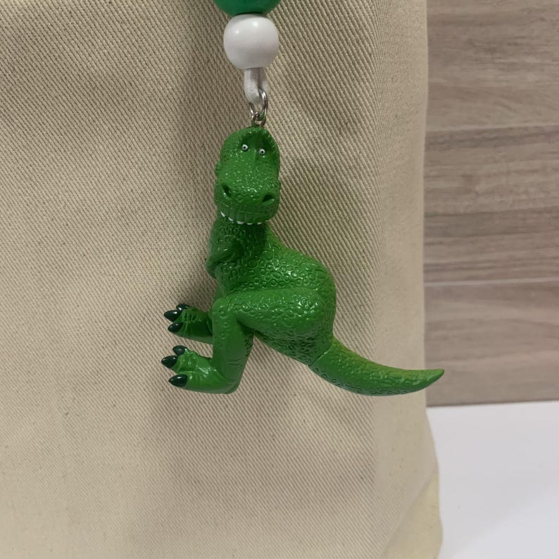 K&ouml;nnte beinhalten: Ein gr&uuml;ner Dinosaurier-Anh&auml;nger mit strukturierter Oberfl&auml;che, der an einer wei&szlig;en und gr&uuml;nen Perlenkette h&auml;ngt. Der Dinosaurier hat eine leuchtend gr&uuml;ne Farbe und schwarze Krallen. Der Anh&auml;nger ist an einem beigefarbenen Stoff befestigt.