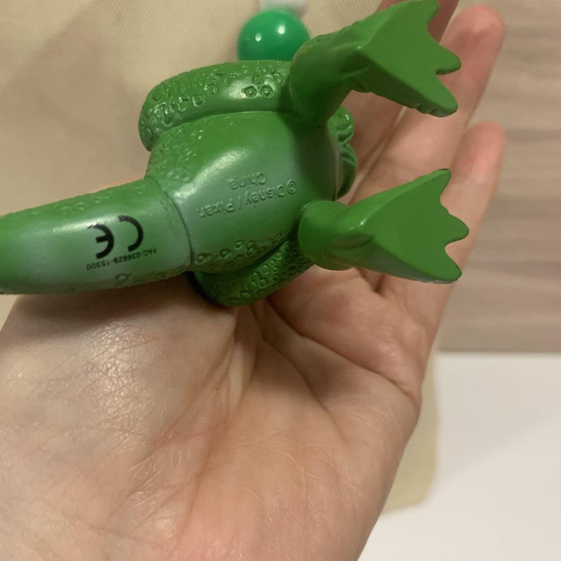 K&ouml;nnte beinhalten: Eine gr&uuml;ne Spielfigur einer Zeichentrickfigur mit strukturiertem K&ouml;rper und gro&szlig;en F&uuml;&szlig;en. Die Figur liegt auf dem R&uuml;cken, wobei die Unterseite sichtbar ist, einschlie&szlig;lich des Textes "&copy;Disney/Pixar China".