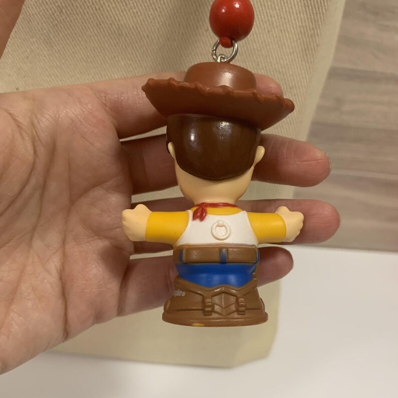 Puede incluir: Un colgante de personaje de Woody con sombrero de vaquero marr&oacute;n, camisa amarilla y pantalones azules. El colgante est&aacute; unido a un collar de cuentas rojas. El juguete es una versi&oacute;n en miniatura del personaje de la pel&iacute;cula Toy Story.