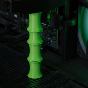 Puede incluir: Un soporte de componente de computadora verde brillante con forma de bambú. El soporte es cilíndrico con secciones segmentadas y está posicionado dentro de una caja de computadora cerca de otros componentes. El fondo incluye un ventilador y otro hardware de computadora.