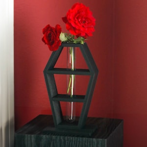 May include: A black wooden coffin-shaped vase with a glass test tube for holding red roses. The vase is sitting on a black wooden pedestal against a red wall.