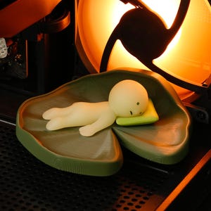 May include: A small, cream-coloured figurine of a person lying on a green lily pad with a yellow pillow. The figurine is resting inside a computer case, near a fan with an orange glow.