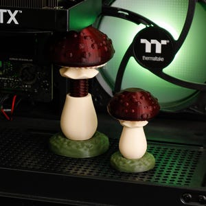 May include: Two 3D-printed mushroom sculptures featuring dark red caps with raised spots, cream-colored stems, and textured green bases. The taller mushroom has a visible threaded stem design, while the shorter one has a smooth, solid stem structure.