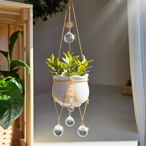 Könnte beinhalten: Ein weißer Keramik-Hängepflanzentopf mit goldener Kette und Kristallakzenten. Der Pflanzentopf enthält eine kleine grüne Pflanze. Die goldene Kette ist an einem dekorativen Sonnenstrahl und einem Kristallkugel aufgehängt. Drei weitere Kristalltropfen hängen darunter.
