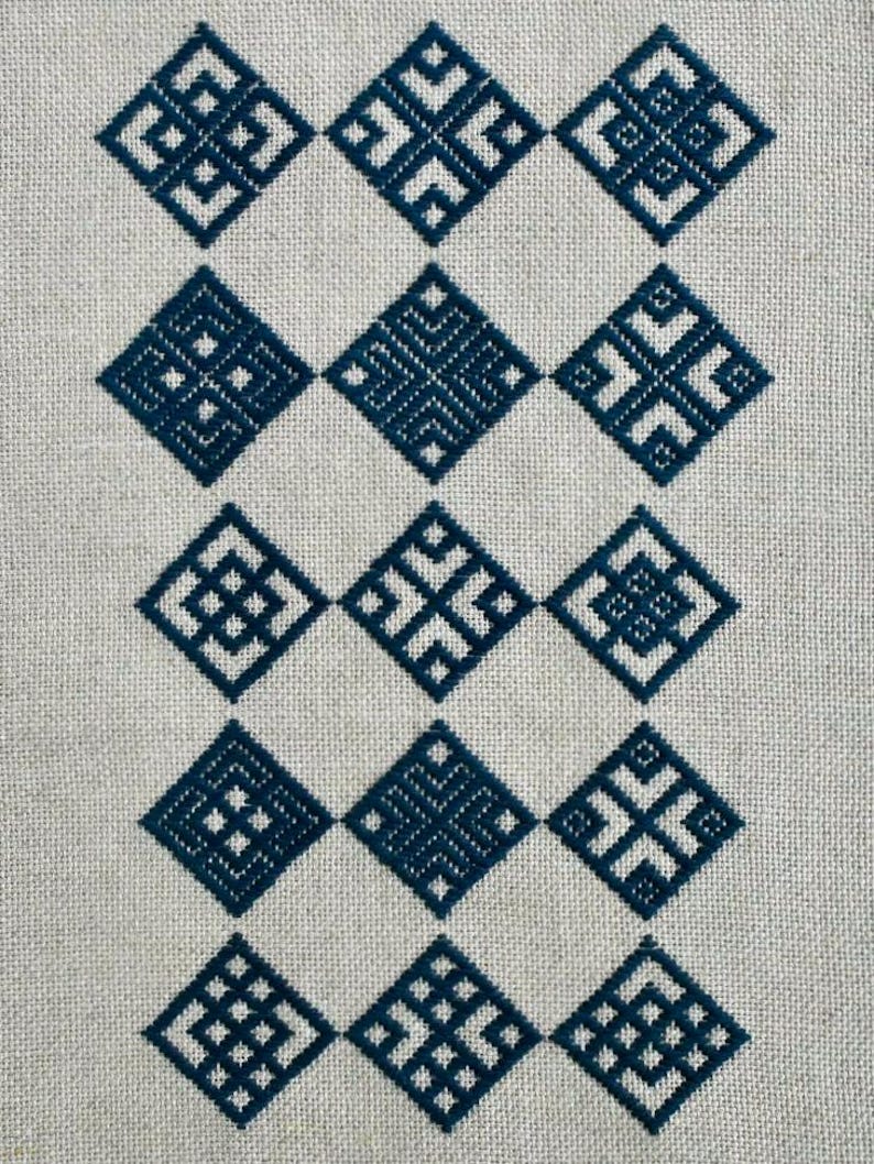 May include: A piece of light beige linen fabric featuring fifteen dark blue embroidered diamond-shaped motifs arranged in a grid of five rows and three columns. Each diamond contains a unique geometric pattern made of straight stitched lines.