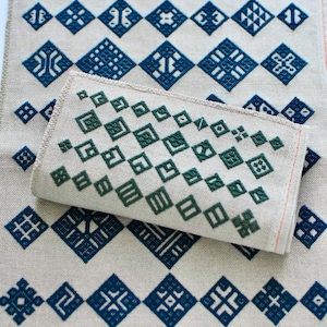 May include: A rectangular piece of light beige fabric featuring a repeating pattern of dark blue geometric diamond shapes and intricate symbols. A smaller, folded piece of the same fabric with dark green diamond patterns rests on top.